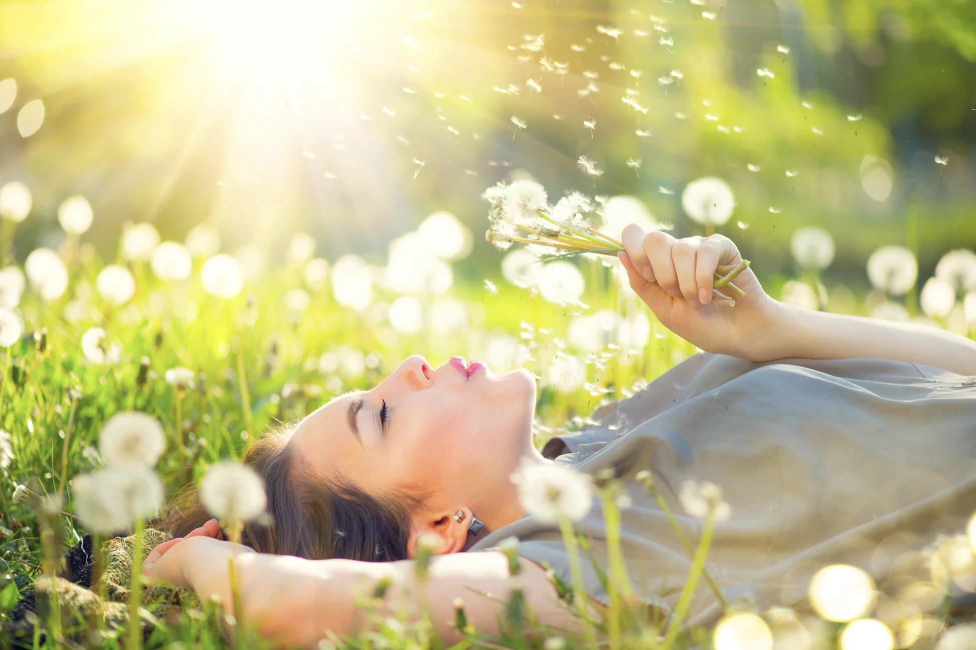 Blog A woman is laying in the grass surrounded by cherished dandelion seeds.