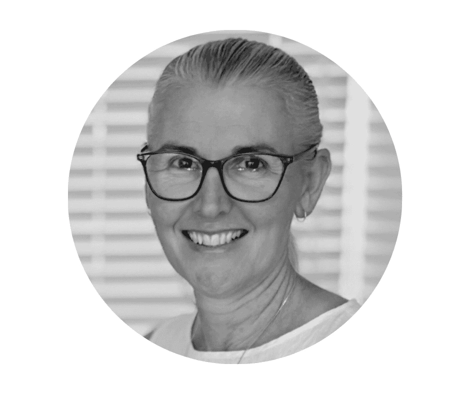 Meet our Babysitters Black and white portrait of a smiling Cherished Cherubs Babysitter with glasses and pulled-back hair, wearing a light top, in front of a background with horizontal blinds.
