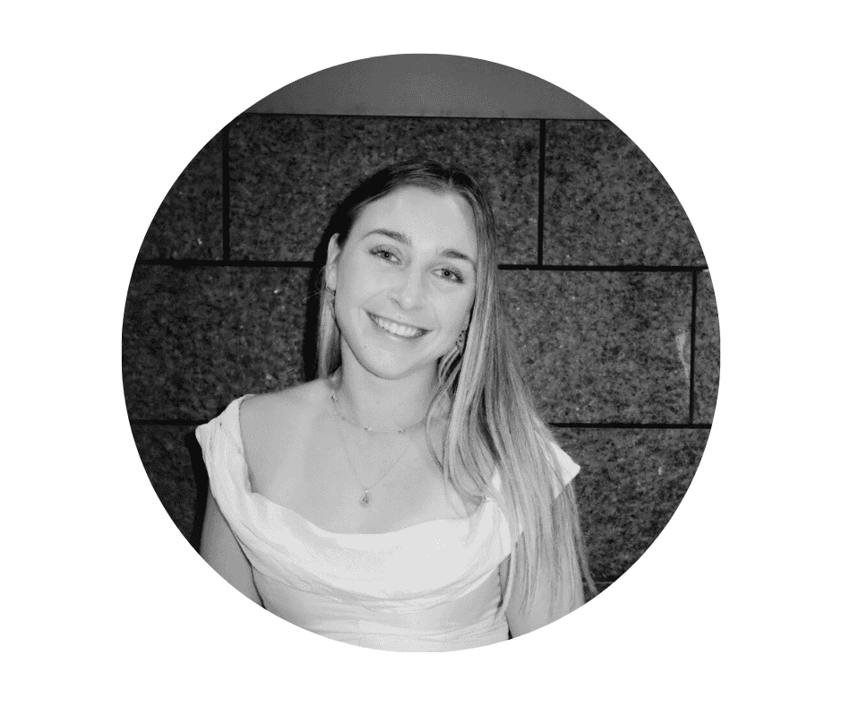 Meet our Babysitters A Cherished Cherubs Babysitter with long hair smiles at the camera, wearing a white top and necklace, standing in front of a tiled stone wall. This black-and-white portrait captures one of our Cherished Cherubs in a timeless moment.