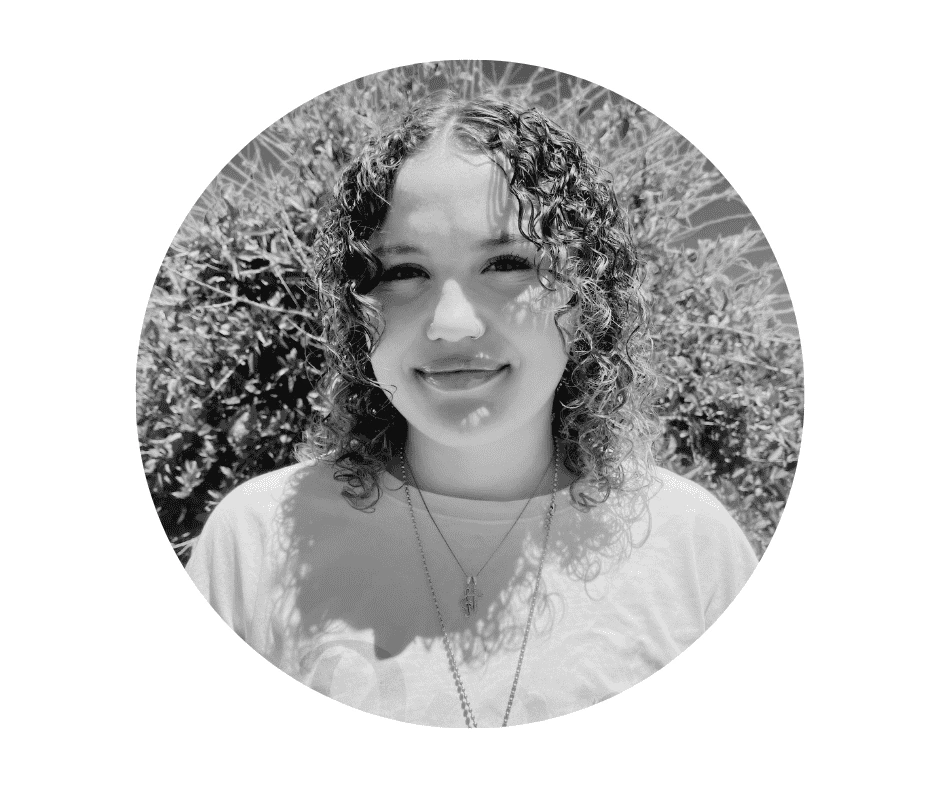 A Cherished Cherubs Babysitter with curly hair, wearing a light-colored shirt and necklaces, stands outdoors in front of foliage, photographed in black and white.