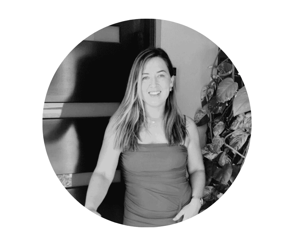 Meet our Babysitters A Cherished Cherubs Babysitter with long hair, wearing a sleeveless dress, stands indoors near a door and a leafy plant, smiling at the camera. This black and white photo captures a moment of joy, echoing the timeless charm of Cherished Cherubs.