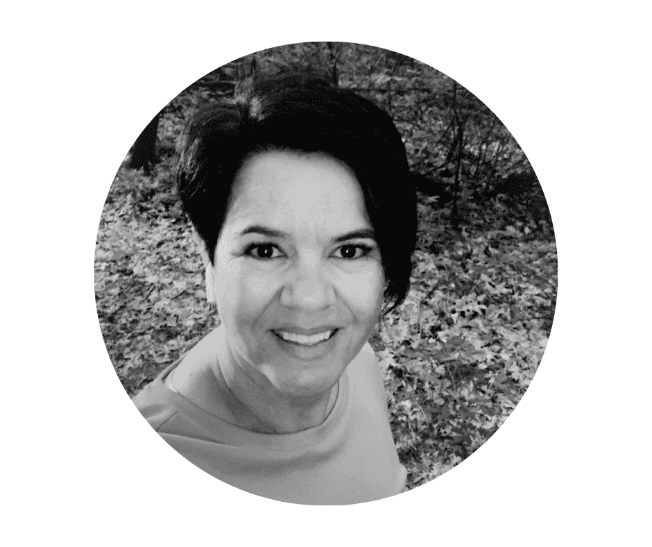 Meet our Babysitters A Cherished Cherubs Babysitter with short dark hair smiles at the camera in an outdoor setting with trees and fallen leaves behind her. The black and white image, cropped in a circular frame, radiates the warmth of a Cherished Cherubs moment.